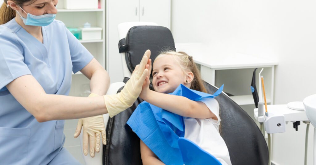 Pediatric dentist high fiving young girl sitting in dentist’s chair.