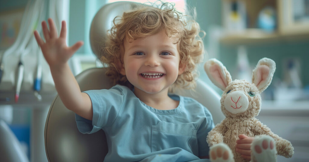 Young boy sitting in dentist’s chair with stuffed rabbit and waving.