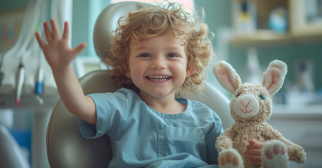 Young boy sitting in dentist’s chair with stuffed rabbit and waving.