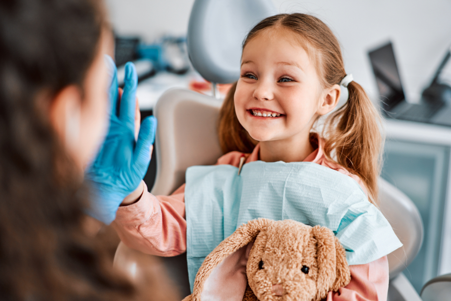 Young girl in dentist’s chair with stuffed bunny hi-fiving dentist.