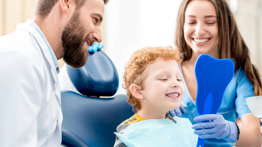 Boy in dentist’s chair smiling at mirror held up by a dentists assistant while a dentist looks on.