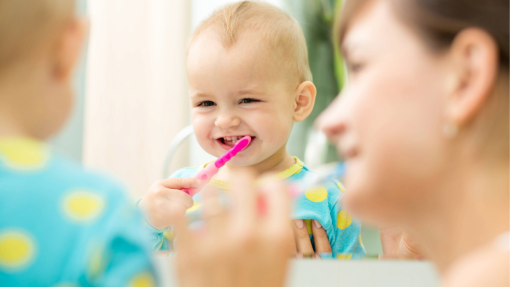 Baby smiling in mirror while mother brushes his teeth.