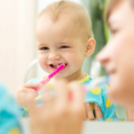 Baby smiling in mirror while mother brushes his teeth.